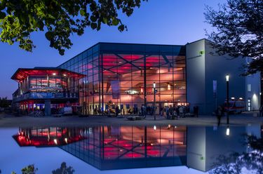 Die OsnabrückHalle, ein Veranstaltungszentrum, erleuchtet in den Abendstunden und sich im Wasser widerspiegelnd.