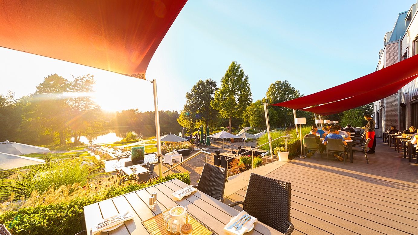 Die Terrasse des See- und Sporthotel Ankum mit Seeblick ist mit roten Sonnensegeln überspannt. Im Hintergrund geht die Sonne unter.